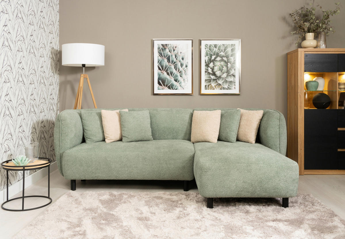 Lomoco ECKSOFA Uma Grün Chenille, links davon: Hochflorteppich in weiß, Beistelltisch in schwarz, rechts davon: Regal, Topfpflanze in hellem Wohnzimmer.