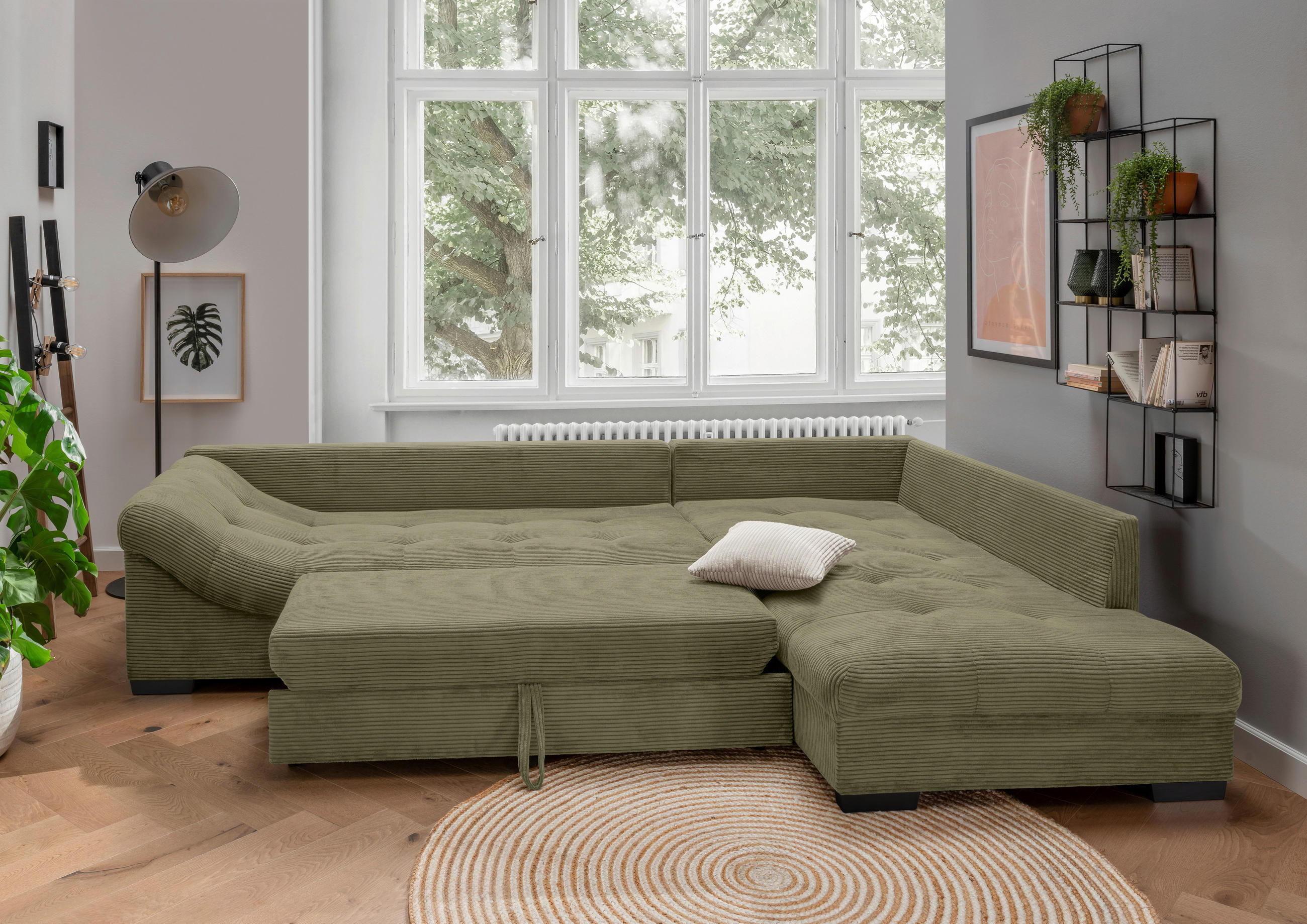 MID.YOU ECKSOFA Olivgrün Cord, links davon: Topfpflanze in grün, rechts davon: Fußmatte, Kissen, Regal in hellem Wohnzimmer.