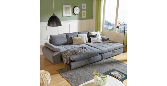 Das Bild zeigt ein modernes Wohnzimmer mit einem grauen Sofa, einem Couchtisch, einem Teppich, einer Stehlampe und einem Vorhang. Auf dem Sofa liegen zwei Kissen mit der Aufschrift 