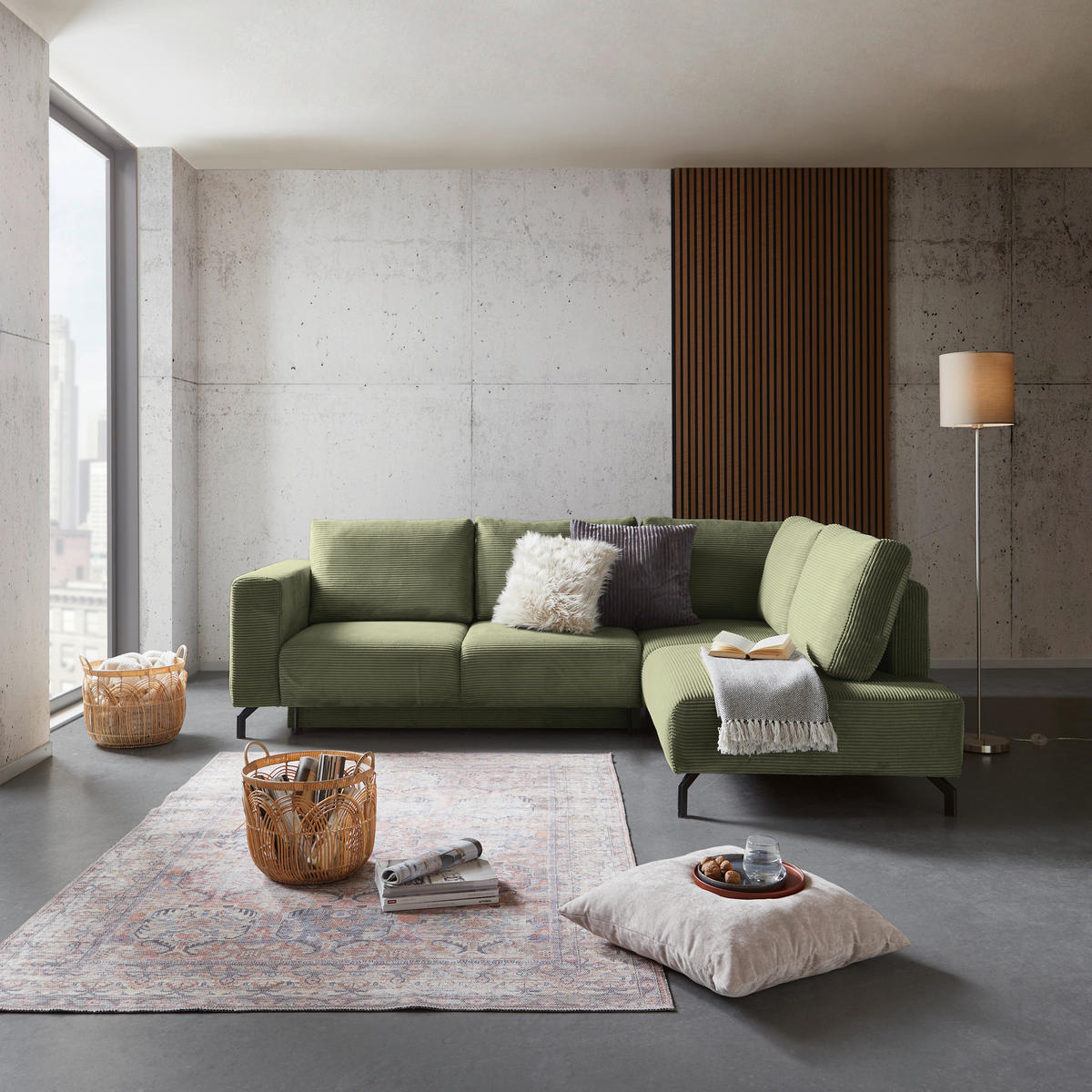 P & B ECKSOFA Grün Cord, links davon: Hochflorteppich, rechts davon: Kissen in weiß, Lektüre, Stehleuchte in hellem Wohnzimmer.