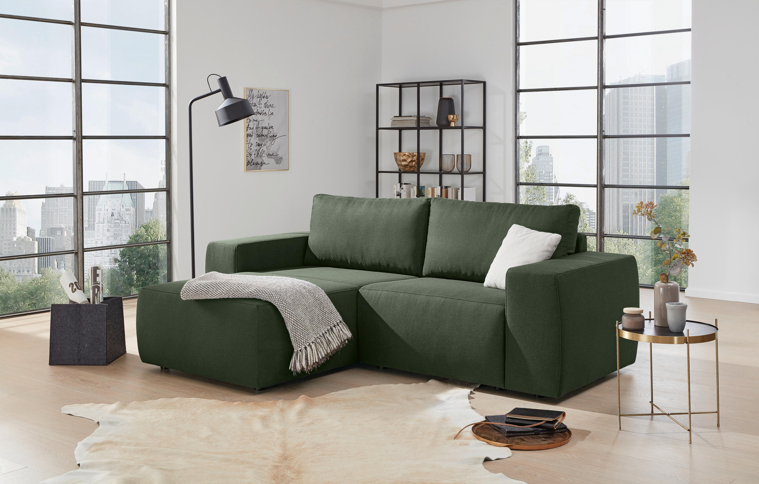 LOOKS by W.Joop ECKSOFA LOOKS II Grün Webstoff, links davon: Kunstfell in grün, rechts davon: Regal in schwarz, Beistelltisch, Lektüre in hellem Wohnzimmer.